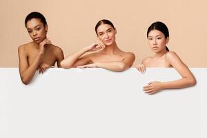 Three Beautiful Diverse Girls Posing Standing Behind White Poster For Text On Gray Studio Background. Free Space photo
