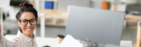 Happy businesswoman having call or taking selfie while working with documents in office and smiling at camera. Confident lady entrepreneur communicating online at workplace, copy space photo