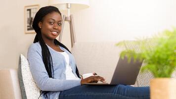 Freelance Concept. Happy Afro Girl Working Online On Laptop And Drinking Coffee, Sitting On Comfortable Sofa At Home photo