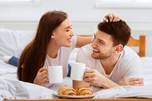 Breakfast In Bed. Happy Couple Having Coffee And Croissants Enjoying Weekend Lying In Bedroom At Home. photo