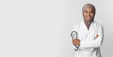 Portrait Of Young Afro Muslim Female Doctor In Hijab Wearing White Coat And Holding Stethoscope, Posing With Folded Arms Over Light Background photo