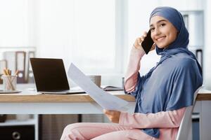 Smiling saudi businesswoman in hijab working with documents and talking on cellphone at workplace in office, side view with empy space photo
