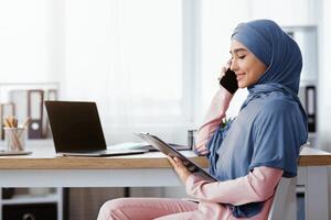 Muslim business lady in hijab holding clipboard, checking documentation and talking on mobile phone with partner while sitting at workplace in office photo