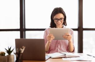 Surfing The Net. Happy young secretary in eyeglasses using digital tablet and smiling, sitting at desk in modern office photo