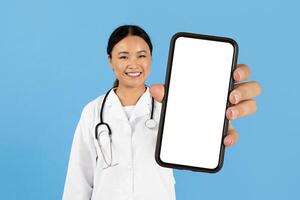 A healthcare worker Asian lady stands against a blue background, smiling warmly while holding up a smartphone with a blank screen, symbolizing telemedicine or digital health options available today. photo