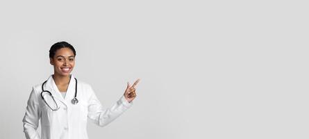 Young black female doctor pointing at copy space on light background, wearing white coat and stethoscope and smiling at camera photo