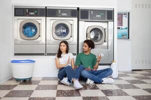 A couple sits on the floor of a laundromat, appearing engaged in conversation. The modern facility features several washing machines nearby, creating a casual atmosphere. photo