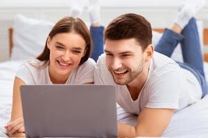 Weekend. Millennial Couple Using Laptop Lying In Bed Watching Movie Spending Weekend At Home. Selective Focus photo