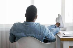 Idle time concept. Rear view of lazy african american employee sitting with his legs on work desk and looking to window, copy space photo