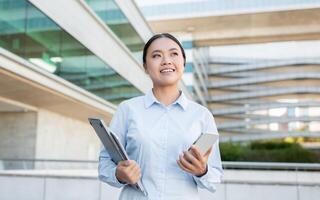 A young professional Asian lady is outdoors in a modern urban setting, smiling confidently while holding a tablet and smartphone. Buildings and greenery add context to her upbeat demeanor. photo