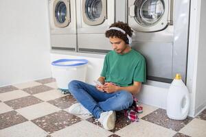 A young individual sits on the floor of a laundromat, wearing headphones and looking at a phone. They are relaxed and engaged while laundry machines operate in the background. photo