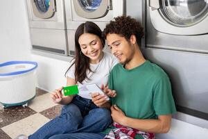 A young couple shares a moment at a laundromat, smiling as they check their smartphone while one holds a credit card. The atmosphere is relaxed and casual during a chore. photo