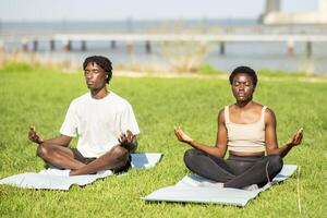 In a peaceful green area by the water, two African American friends sit cross-legged on mats, eyes closed, engaged in meditation. The calm atmosphere enhances their focus and relaxation. photo