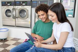 Two young adults sit together in a laundromat, smiling as they look at a tablet. The laundry machines are in the background, indicating a relaxed atmosphere while they wait. photo
