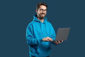 A young man smiles while using a laptop, dressed in a blue hoodie. He stands against a solid background, showcasing a relaxed and engaged attitude while working. photo