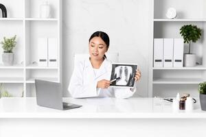 A medical professional Asian lady in a white coat discusses a chest X-ray with a patient during a consultation. The doctor highlights important areas on the X-ray while seated at a desk. photo