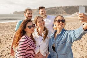 A group of five friends smiles happily as they take a selfie on a sandy beach. The sun sets in the background, casting golden light over the cheerful gathering. photo