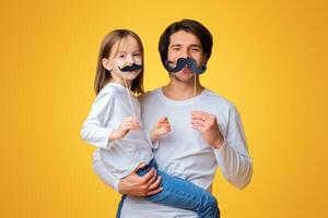 Funny little girl and daddy have mustaches on sticks, posing over yellow background photo