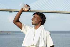 A cheerful black man with dreadlocks stands by the river, enjoying a refreshing drink from a water bottle while a large bridge stretches across the background. His smile reflects his satisfaction. photo