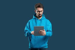 A young man wearing a blue hoodie focuses on a tablet with a serious expression, standing against a solid dark background. He appears to be engaged in thoughtful activity with the device. photo