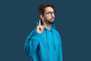 un joven hombre soportes adentro vistiendo un brillante azul sudadera, levantamiento su índice dedo como Si a señal un pensamiento o idea. él muestra un pensativo expresión en contra un profundo azul antecedentes. foto