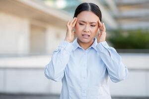 A young Asian woman is standing outside in an urban environment, holding her head with a look of discomfort, likely dealing with a headache or stress, while dressed in a professional outfit. photo