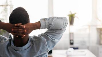 Bored at work. Rear view of african american employee sitting at workplace with hands behind his head, looking to window in modern office, photo