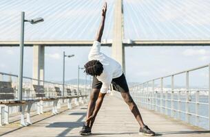 African American young man performs a side stretch on a wooden platform located by a river, enjoying the sunny weather and a view of a modern bridge in the background. photo