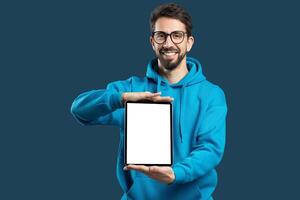 A young man wearing a blue hoodie smiles while holding a blank tablet in front of him. The background is a solid color, emphasizing his expression and the device he showcases. photo