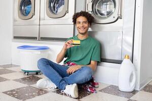 A young man sits on the floor of a laundromat, smiling as he holds a laundry card in his hand. Washing machines are visible in the background, creating a casual atmosphere. photo