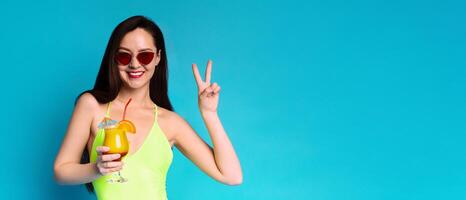 A woman in a yellow swimsuit is smiling and holding a cocktail while making a peace sign. She is wearing sunglasses and is standing in front of a bright blue background, copy space photo