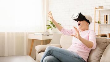 Old woman is seated on a couch, wearing a virtual reality headset. She appears engaged in an immersive digital experience, with her hands resting on her lap. photo