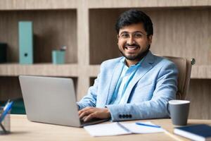 A young Indian man in a blue suit sits at a desk in an office, smiling as he types on a laptop computer. He has a cup of coffee beside him, and there are papers and a notepad on the desk. photo