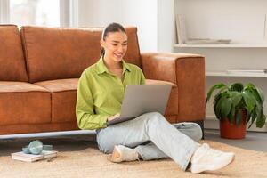 A woman is sitting on the floor, leaning against a wall, and using a laptop. She is focused on the screen, typing and scrolling through information. photo