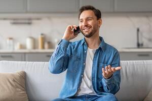 A man with a beard is sitting on a white couch in his home while talking on his phone. He is wearing a blue denim shirt and jeans. He is smiling and gesturing with his hand as he speaks. photo