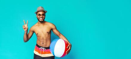 A smiling black man with a beard wearing sunglasses and a hat holds a beach ball while standing in front of a solid blue background. He is wearing colorful swim trunks and making a peace sign photo