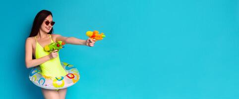 A woman in a bright yellow swimsuit and sunglasses holds two water guns, one in each hand. She has a playful expression on her face and is looking towards the right side of the frame photo