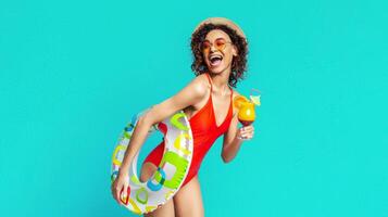 African American woman in a red swimsuit and a straw hat is holding a cocktail and an inflatable ring. She is smiling and appears to be in a playful mood. The background is a solid blue color. photo