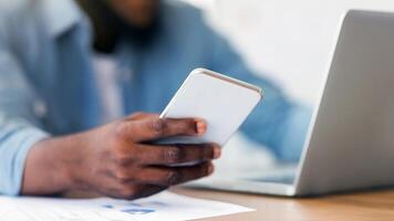 Devices for business. Unrecognizable black entrepreneur using smartphone and working on laptop in modern office, selective focus on phone photo