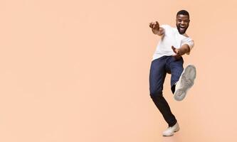 A young Black man in a white t-shirt and blue jeans is dancing in front of a peach-colored background. He has a wide smile on his face and is lifting his right leg, copy space photo