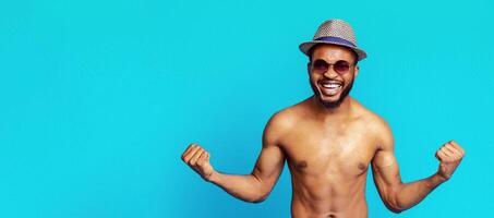 A shirtless African American man with a beard is wearing a straw hat and sunglasses. He is smiling broadly and flexing both arms. The man is standing in front of a bright blue background. photo