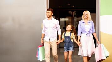 A family, consisting of a father, mother, and young daughter, are walking out of a shopping center. They are holding hands and carrying colorful shopping bags, copy space photo