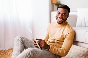 Black Guy Using Tablet Computer Smiling At Camera Sitting On Floor At Home. Selective Focus photo
