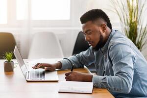African American employee using laptop and taking notes while working in office, side view with copy space photo