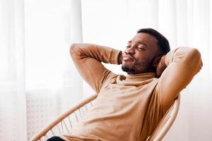 Weekend At Home. Lazy Black Guy Relaxing Sitting On Modern Chair Against Window Indoor. Selective Focus photo