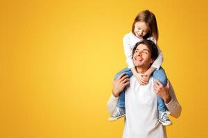 Happy father carrying his lovely cheerful little daughter on shoulders, having fun together over yellow background, copy space photo