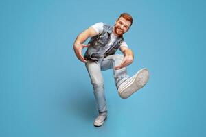 Come on. Smiling millennial caucasian guy fool around and showing his shoe sole at camera, full length, blue studio background photo