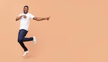 A happy African American young man is jumping in the air while pointing to the right with a big smile on his face. He is wearing a white t-shirt and dark blue jeans, panorama with copy space photo