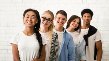 A cheerful group of two girls and three guys are lined up shoulder to shoulder, posing casually against a textured white brick wall. photo