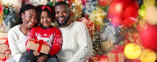 Joyful afro family of three posing with gift over Christmas tree background, smiling and looking at camera, closeup photo